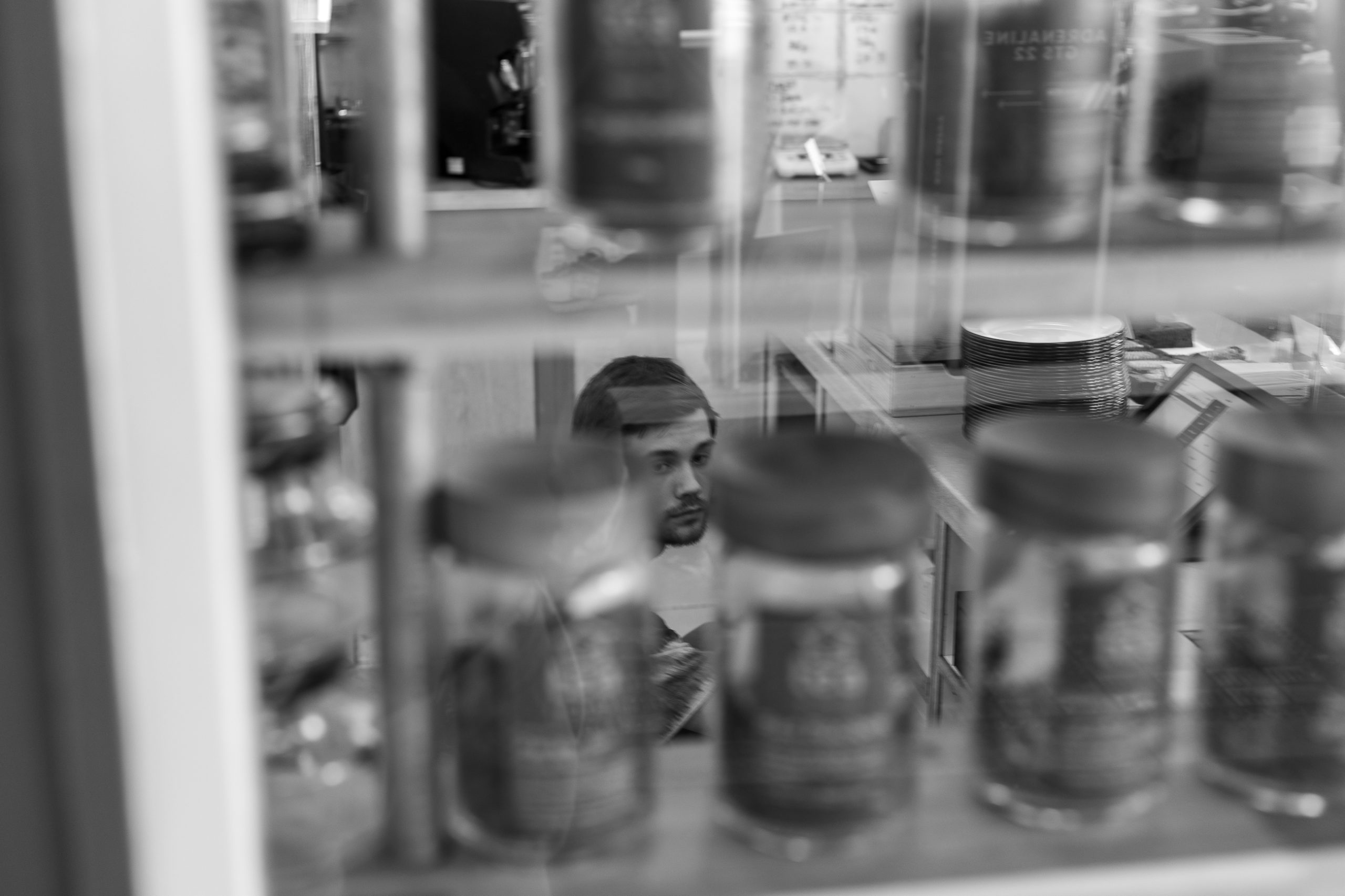 A presenting male crouches behind a counter. The picture is shot through a window and glass jars sitting on shelves.