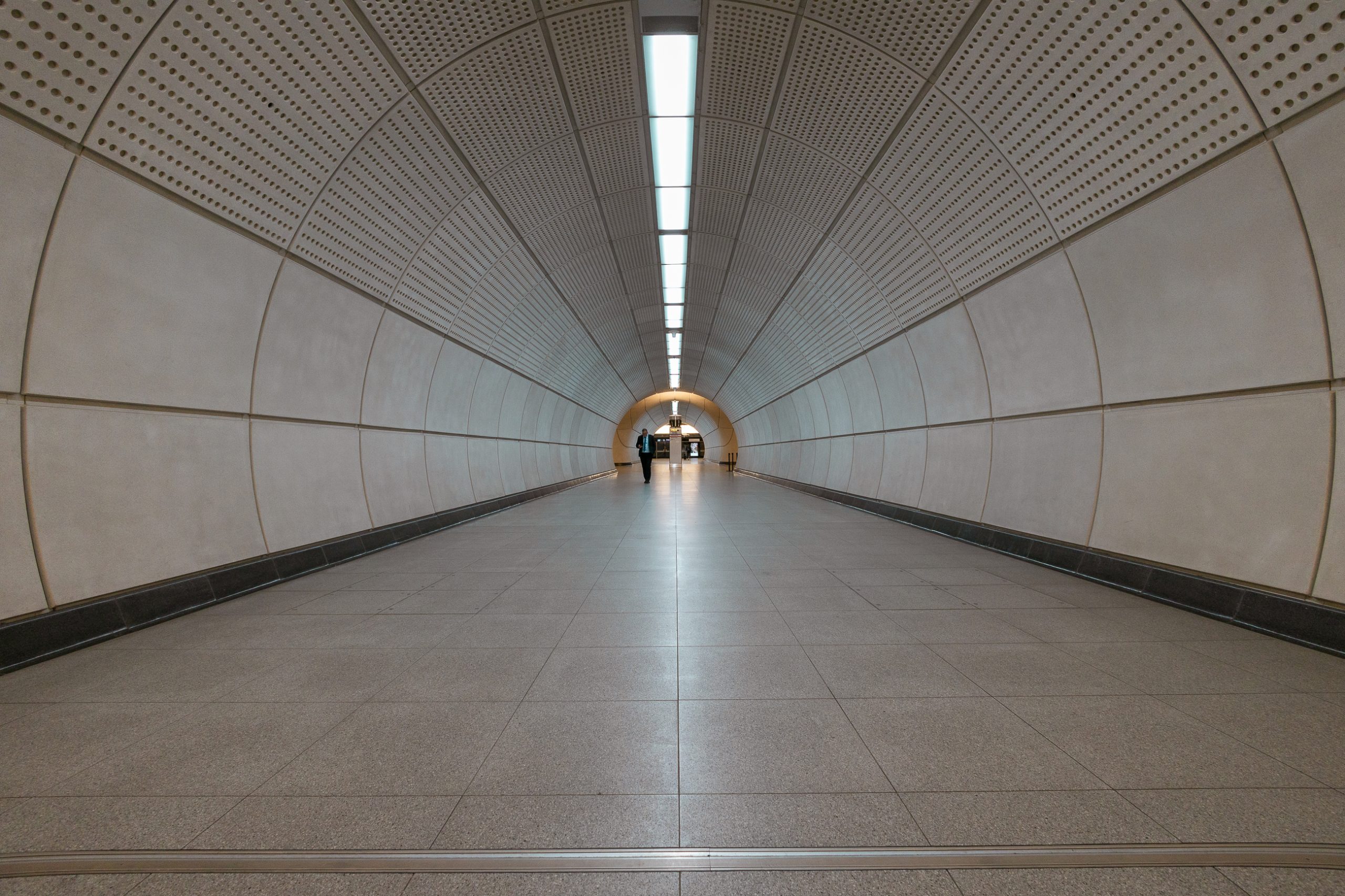 An image taken below standing waist height of a connecting tunnel between the Elizabeth Line platforms at Tottenham Court Road station. In the distance, a male figure approaches.
