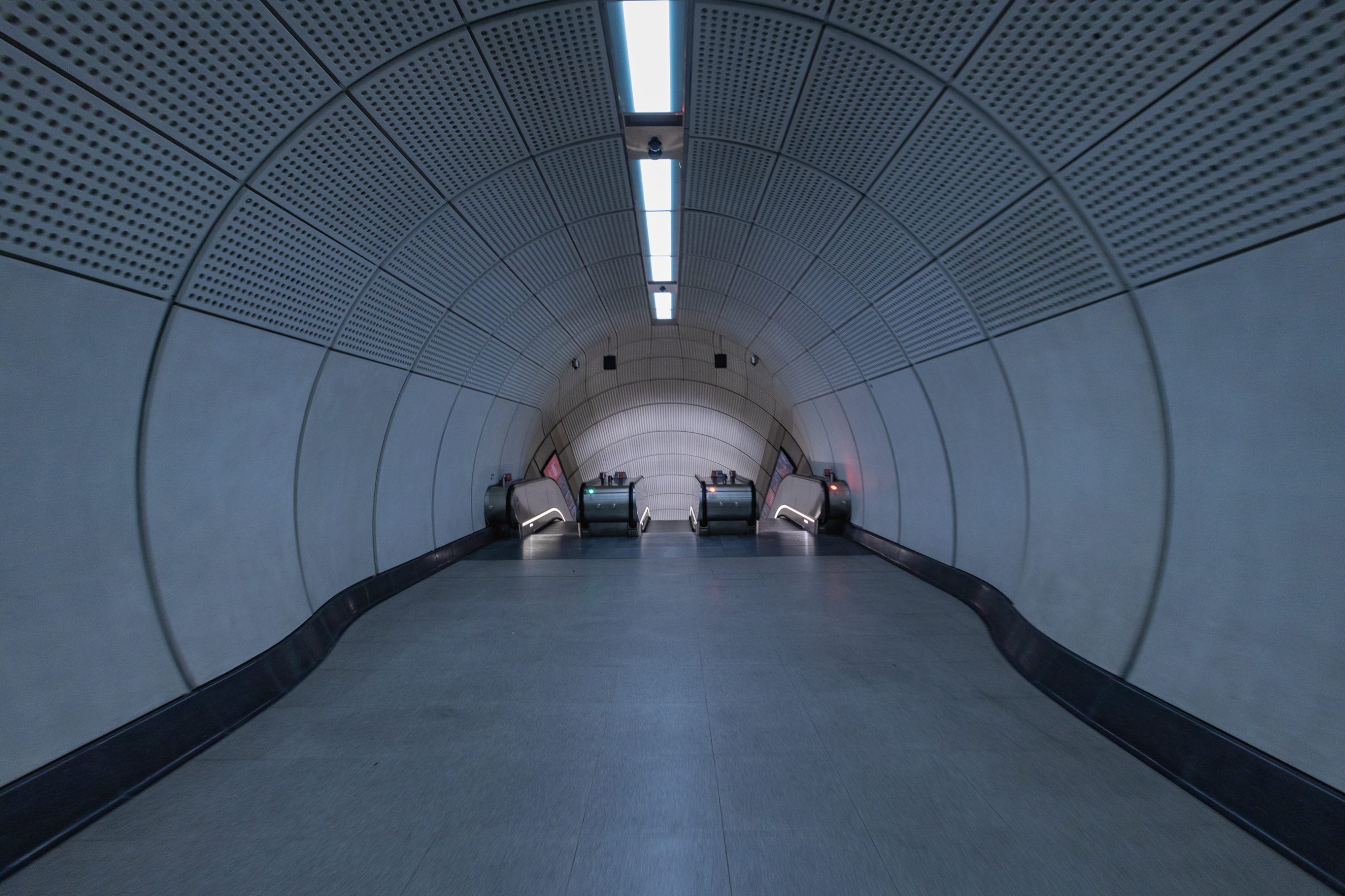 A standing height picture of the end of a tunnel leading to three escalators. The tunnel widens, producing an optical illusion of the floor rising in a slightly disturbing way