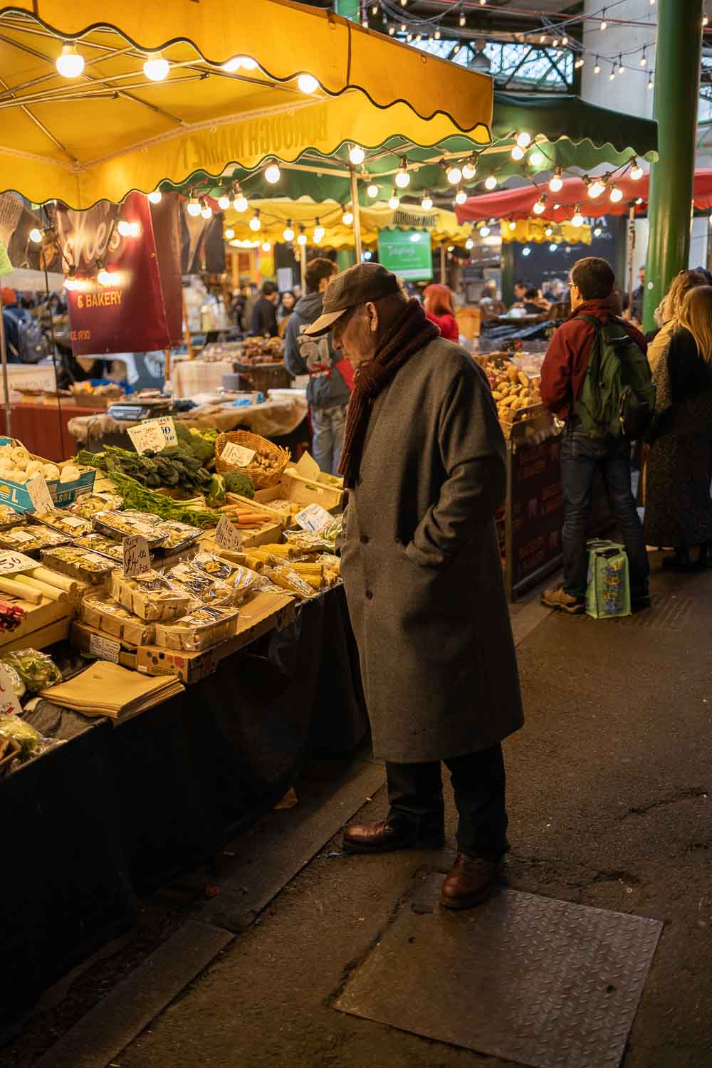 check. A tourist? A regular? Either way, I think they know what they're looking for and this isn't it. A person in an overcoat, baseball cap and polished brown shoes leans slightly forwards as they inspect some groceries for sale