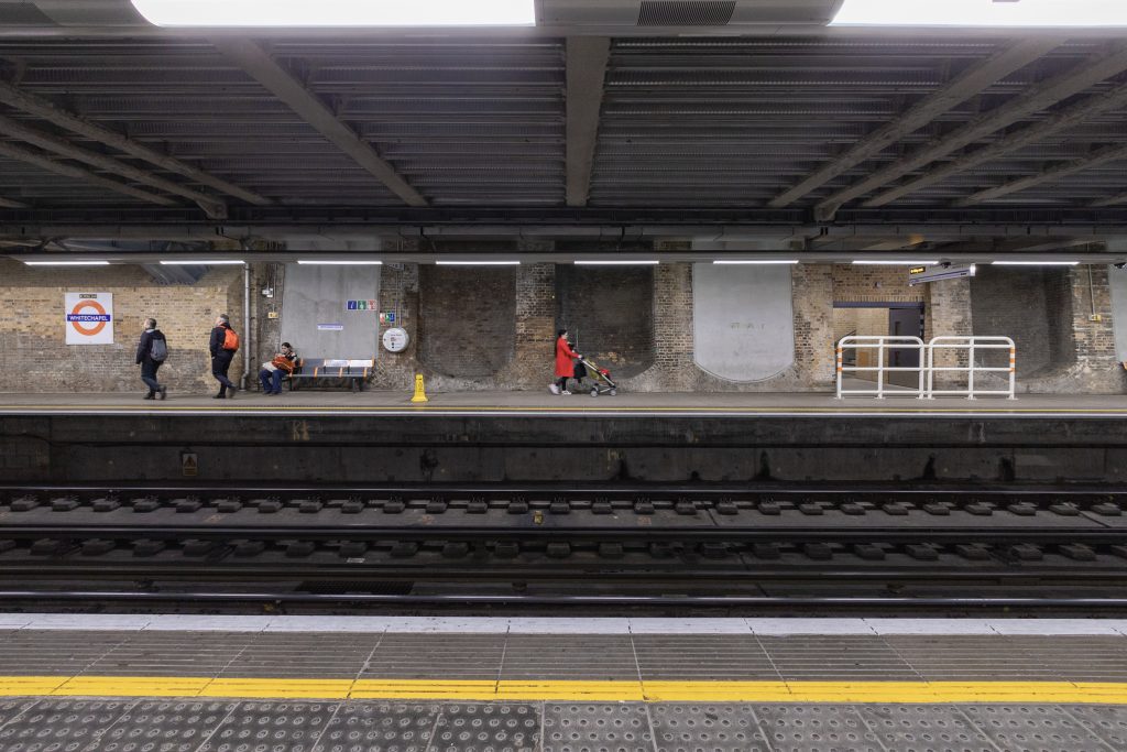 A woman wearing a red coat and pushing a stroller walk along the Eastbound platform of the District Line of London's Undeground transit system at Whitechapel
