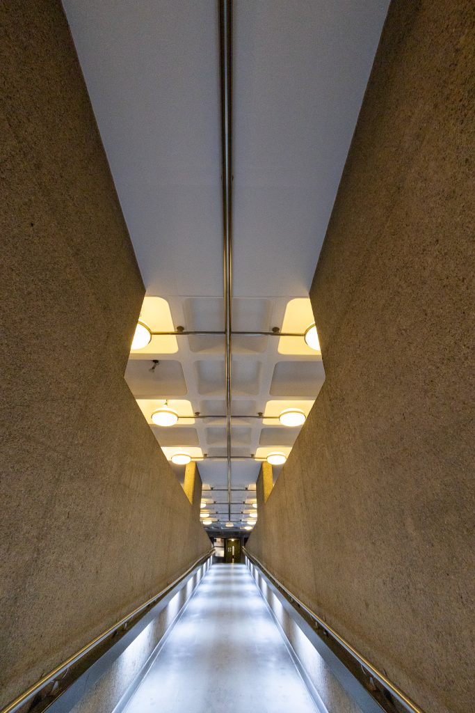A photo of an upward-sloping internal corridor at the Barbican Centre showing the ceiling and handrail lighting